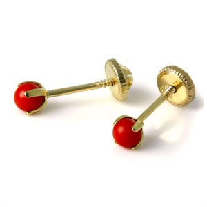 May include: A pair of gold stud earrings with a bright red, round gemstone. The earrings have a screw-back closure and are set in a gold bezel. The earrings are displayed on a white background.