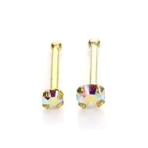 Pode incluir: Dois piercings de nariz dourados com cristais quadrados iridescentes. Cada piercing apresenta um pequeno poste redondo e uma moldura decorativa em torno do cristal. Os cristais refletem um espectro de cores.
