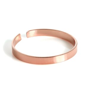 Può includere: Un bracciale rigido in metallo color oro rosa con una finitura liscia e lucida. Il bracciale presenta un design circolare semplice e aperto con un profilo a fascia piatta, mostrando una lucentezza metallica uniforme su tutta la superficie.