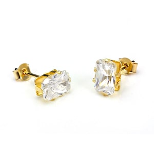 May include: A pair of gold-colored stud earrings, each featuring a rectangular, clear gemstone. The earrings are set in a gold-toned metal with a post and butterfly backing. The gemstones are faceted, reflecting light.