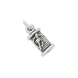 May include: A small, three-dimensional silver charm shaped like a spool of thread. The metal piece features detailed horizontal grooves representing wound thread and a small loop at the top for attachment, finished with a polished silver sheen.
