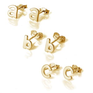May include: Gold-coloured stud earrings shaped as the letters 'a', 'b', and 'c'. The earrings are presented on a white surface, each letter featuring a post for ear insertion. The letters are in a lowercase, sans-serif font.
