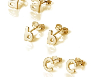 Puede incluir: Pendientes dorados con forma de las letras 'a', 'b' y 'c'. Los pendientes se muestran sobre una superficie blanca, cada letra tiene un poste para insertar en la oreja. Las letras son en minúsculas, sin serifa.