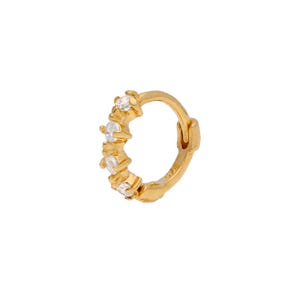 May include: A gold-coloured hoop earring featuring five clear, round gemstones. The earring has a hinged closure. The gemstones are evenly spaced along the front of the hoop. The background is white.