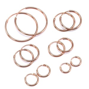 Peut inclure: Plusieurs paires de boucles d'oreilles créoles en or rose de différentes tailles, sur fond blanc. Les boucles d'oreilles varient des grandes créoles aux modèles plus petits et discrets. Le métal a une teinte rosée chaude, et le design simple les rend polyvalentes.
