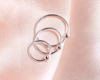 May include: Three silver hoop earrings of varying sizes, each with a small ball accent. The earrings are displayed on a soft, pink fabric, creating a delicate and elegant presentation. The hoops are simple and minimalist in design.