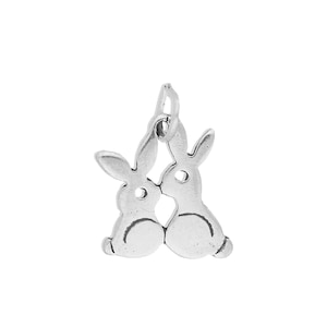 May include: A silver-toned metal charm featuring two rabbits facing each other in a mirrored silhouette. The rabbits are connected at the ears and bodies, with a small circular cutout near the head of each rabbit and a jump ring attached at the top.