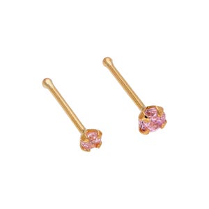 May include: Two gold-coloured nose studs, each featuring a small, round, light pink gemstone. The studs have a simple, elegant design, with a straight post and a decorative setting for the gemstone. The background is plain white.