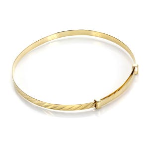 May include: A gold-colored bangle bracelet with a textured, curved design on the front. The bracelet has a clasp closure and is set against a white background. Jewelry.