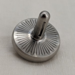 Könnte beinhalten: Eine silberne Metallkreisel mit einem dekorativen Muster auf der Oberseite. Der Kreisel hat eine erhöhte Mitte mit einer Rille.