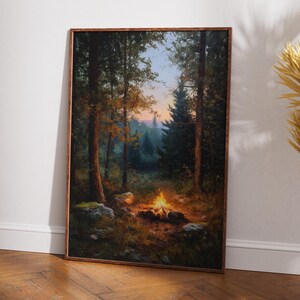 May include: A framed landscape painting depicts a campfire scene in a forest. The artwork features tall trees, a glowing fire, and a soft, colorful sky. The painting is set in a wooden frame and leans against a white wall.
