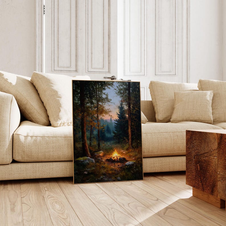 May include: A framed landscape painting depicts a campfire scene in a forest. The artwork features tall trees, a glowing fire, and a colorful sunset. The painting is displayed on a wooden floor, leaning against a beige sofa.