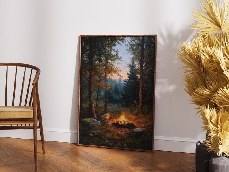 May include: A framed landscape painting depicts a campfire scene in a forest. The artwork features tall trees, a glowing fire, and a soft, colorful sky. The painting is set in a wooden frame and leans against a white wall.