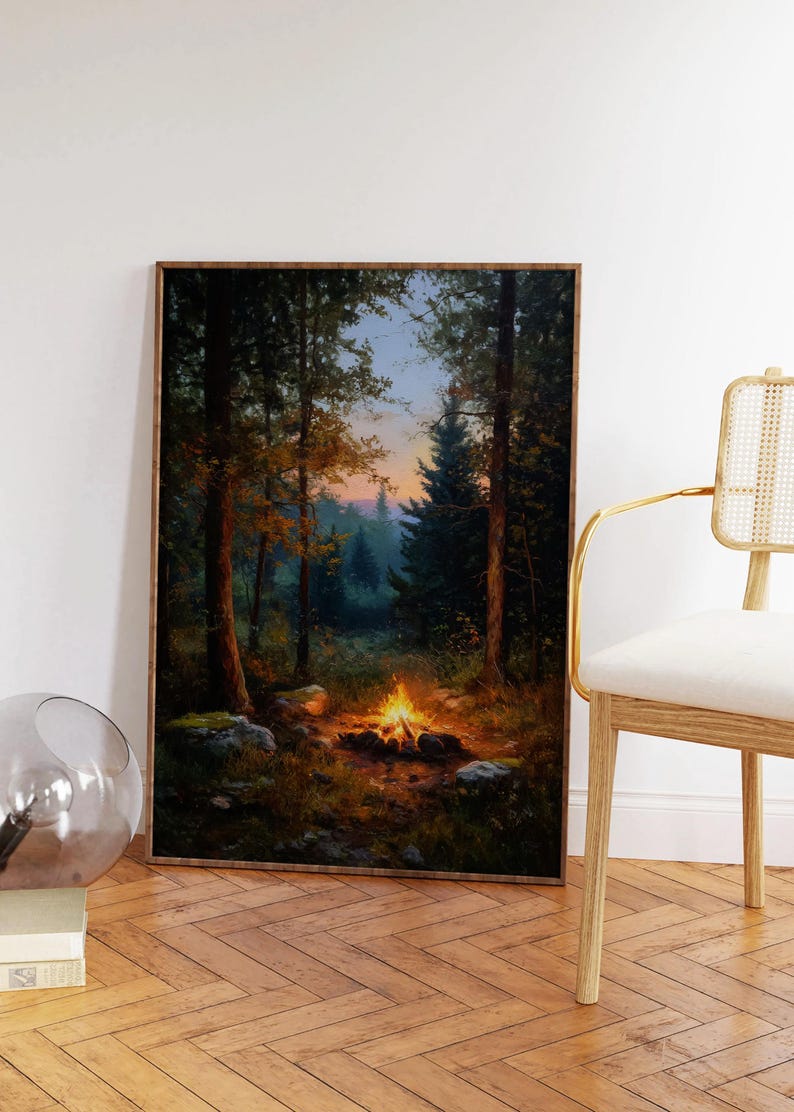 May include: A framed landscape painting depicts a campfire in a forest at dusk. The artwork features tall trees, a glowing fire, and a colorful sky. The painting is displayed on a wooden floor, with a modern chair in the background.