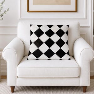 May include: A square throw pillow featuring a high-contrast black and white harlequin diamond pattern. The pillow is centered on a white upholstered armchair with rounded arms and wooden legs, showcasing a geometric design across its surface.