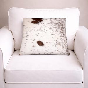 May include: A square throw pillow featuring a cowhide pattern with a white base and irregular dark brown and black spots. The texture appears soft and plush, covering the entire front surface of the pillow in a natural, mottled animal print design.