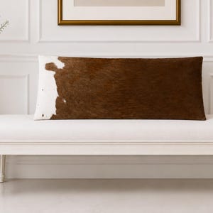 May include: A rectangular lumbar pillow featuring a brown and white cowhide pattern. The pillow has a textured surface with a large brown section covering the majority of the front and a white patch on the left side, creating a distinct contrast.