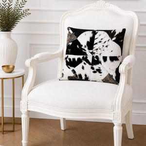 May include: A white wooden armchair with carved detailing and a white upholstered seat. A square throw pillow with a black and white cowhide patchwork pattern sits on the seat. The chair features curved armrests and fluted legs.