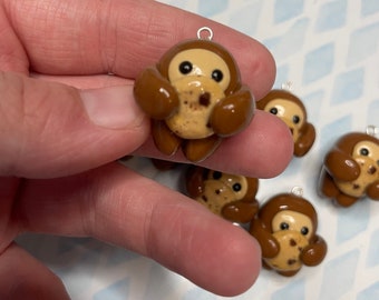 May include: A set of brown clay charms shaped like monkeys holding chocolate chip cookies. The charms have black eyes and a light brown face.