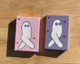May include: Two small canvases with painted ghost figures. The ghost on the pink canvas is facing left, while the ghost on the purple canvas is facing right. Both ghosts have white bodies with black eyes and are surrounded by small pink hearts.