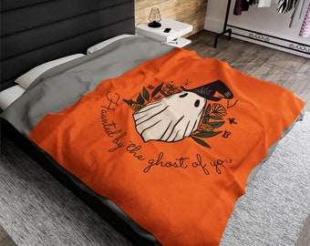 May include: An orange blanket with a ghost illustration wearing a witch hat, surrounded by flowers and leaves. The text "Haunted by the ghost of you" is written in a cursive font. The blanket is on a bed with gray sheets.