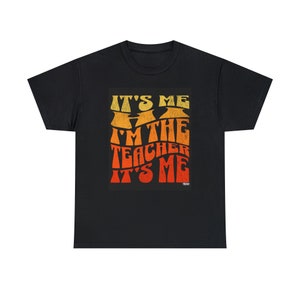 May include: Black t-shirt with the text "IT'S ME I'M THE TEACHER IT'S ME" in a retro, orange and yellow font. The design has a distressed, vintage look. A casual, comfortable tee for teachers.