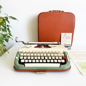 May include: A vintage Princess 300 typewriter in cream and green, with white keys and a red tab key. It is accompanied by a brown carrying case and paperwork. The typewriter has a retro design.