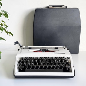 Op de afbeelding: Een witte handmatige typemachine met zwarte ronde toetsen en een zwarte wagenretourhendel, met de merknaam Tippa op de voorzijde gedrukt. Een zwarte harde draagkoffer met een zilverkleurig handvat staat rechtop achter de typemachine.