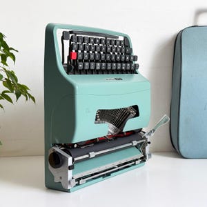May include: A mint green Erika Brillante typewriter positioned vertically, featuring a black keyboard with a single red key, a metal carriage mechanism, and a matching light blue carrying case placed to the right of the machine.