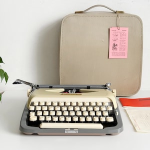 Op de afbeelding: Vintage crème schrijfmachine met een grijze en bruine basis en witte toetsen. Een beige draagtas met handvat en een roze label staat ernaast. De schrijfmachine staat op een wit oppervlak.