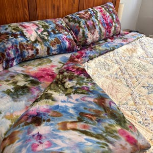 May include: A set of bedding featuring a tie-dye pattern in shades of blue, pink, and olive green. The set includes two rectangular pillowcases and a flat sheet, all displaying a swirling, multi-colored design across the fabric surface.