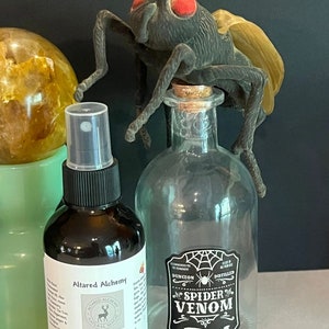 May include: A clear glass bottle with a black and white label that reads "Dungeon Distilled Spider Venom" and a black spider web design. A black plastic fly is perched on top of the bottle. A brown glass bottle with a black and white label that reads "Altared Alchemy Bug Be Gone Spray" is in the foreground.