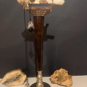 May include: A decorative stand with a bone-like top, a dark brown wooden column, and a silver base. A black cord with a crystal pendant hangs from the top. Two beige rocks are on the base.