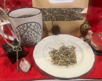 May include: A white mug with a black design that says "Witches Brew Hexy Witch" and a black pentagram. The mug is filled with a brown liquid. There is a white plate with dried herbs on it, a crystal, a key, and a small incense burner.