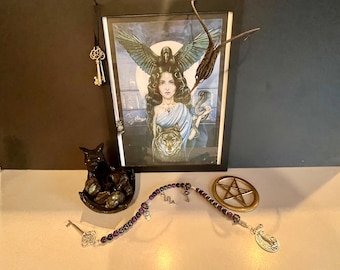 May include: A framed print of a woman with long dark hair, wearing a blue robe, holding a snake and a raven. The print is surrounded by a black frame. A black cat figurine sits in a bowl with stones. A purple beaded necklace with charms, including keys and a snake, is laid out on a white surface. A gold pentagram sits on the surface.