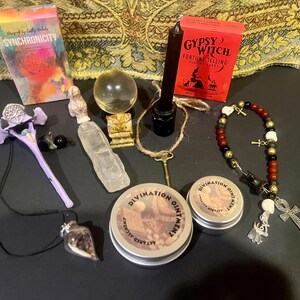 May include: A collection of divination and witchcraft items including a deck of Gypsy Witch Fortune Telling Playing Cards, a crystal ball, a clear quartz crystal, a black candle, a purple wand, a necklace with a skull pendant, a necklace with a crystal pendant, a beaded bracelet with a skull and an ankh pendant, and two tins of divination ointment.
