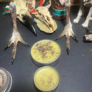 May include: Two small round tins of green balm with a light brown speckled texture. The tins are on a dark gray surface.