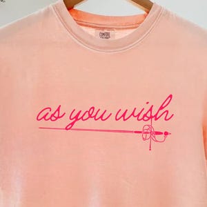 May include: A peach-colored t-shirt with the phrase "as you wish" in pink script, accompanied by a sword illustration. The short-sleeved shirt is hanging on a wooden hanger.