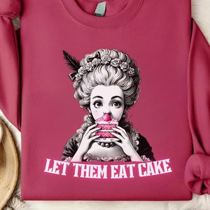 May include: A pink sweatshirt with a graphic of a woman in a white dress and a large white wig eating a pink cake. The text "Let Them Eat Cake" is printed below the graphic.