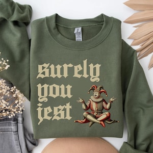 May include: An olive green sweatshirt with the text "Surely You Jest" in a vintage font. The design includes an illustration of a jester in red, white, and brown clothing, with a red and black hat.