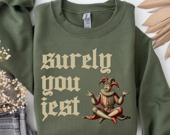 Könnte beinhalten: Ein olivgrünes Sweatshirt mit dem Text "Surely You Jest" in einer Vintage-Schriftart. Das Design zeigt eine Illustration eines sitzenden Narren in roter, weißer und brauner Kleidung mit einem rot-schwarzen Hut.