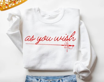 May include: A white crewneck sweatshirt featuring the red text "as you wish" in a cursive script, positioned above a red graphic of a fencing sword. A gold-colored crown with red accents is visible in the upper left corner.