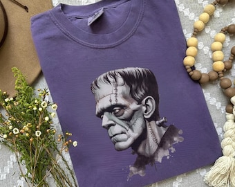 May include: A purple t-shirt with a graphic of Frankenstein's monster. The illustration is in grayscale, showing stitches and a serious expression. The shirt is a solid colour and looks to be made of a soft material, suitable for casual wear.