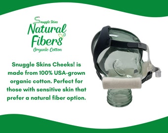 Può includere: Una maschera per dormire in cotone bianco con un cinturino regolabile nero. La maschera è etichettata "Snuggle Skins Cheeks!" e "Natural Fibers Organic Cotton".