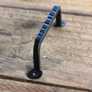 Peut inclure: Poignée de meuble en fer forgé noir avec une poignée texturée. La poignée comporte une série de rainures horizontales, et la poignée est montée sur une surface en bois. La quincaillerie est conçue pour les armoires et les tiroirs.