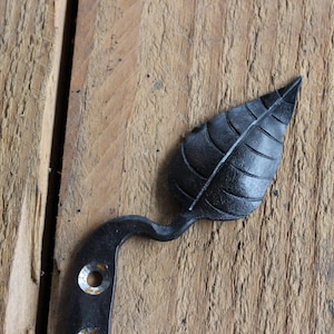 Puede incluir: Bisagra de puerta de metal negro en forma de hoja con dos agujeros para montaje. La bisagra está sobre una superficie de madera.