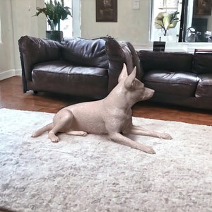 Puede incluir: Una escultura de perro de cer&aacute;mica beige claro tumbada en una alfombra blanca y esponjosa en un sal&oacute;n. El perro est&aacute; en una pose relajada con la cabeza girada hacia un lado.
