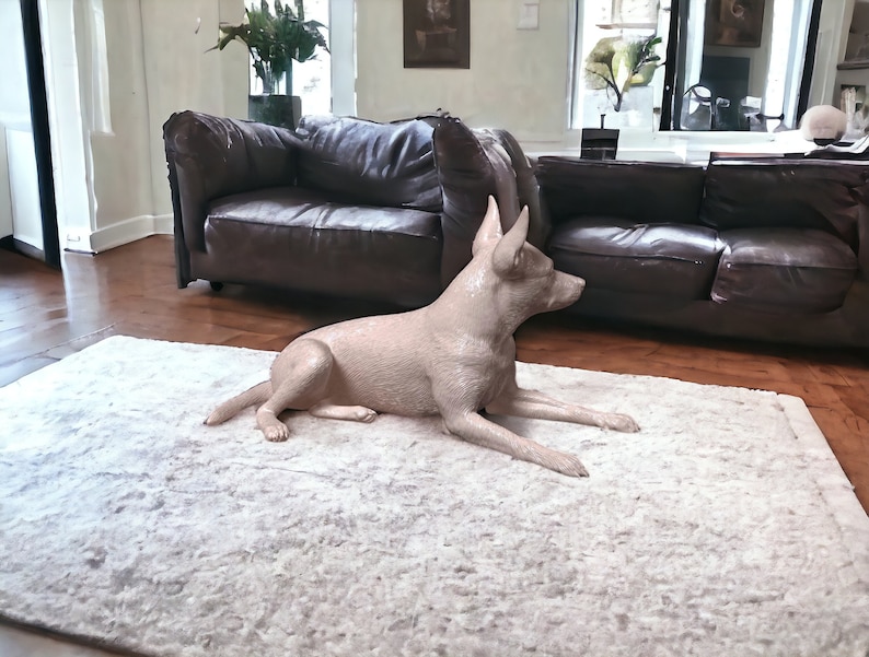 Puede incluir: Una escultura de perro de cer&aacute;mica beige claro tumbada en una alfombra blanca y esponjosa en un sal&oacute;n. El perro est&aacute; en una pose relajada con la cabeza girada hacia un lado.