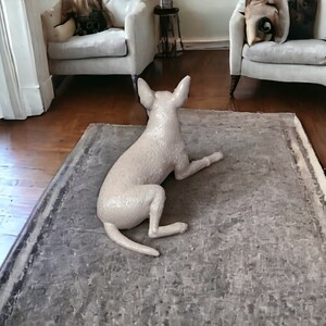 Puede incluir: Una figurita de perro de cer&aacute;mica blanca tumbada de lado sobre una alfombra gris. El perro est&aacute; de espaldas a la c&aacute;mara y tiene la cola enroscada.