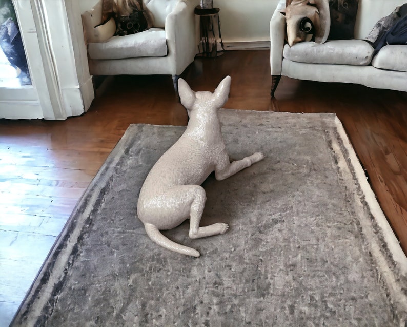 Puede incluir: Una figurita de perro de cer&aacute;mica blanca tumbada de lado sobre una alfombra gris. El perro est&aacute; de espaldas a la c&aacute;mara y tiene la cola enroscada.